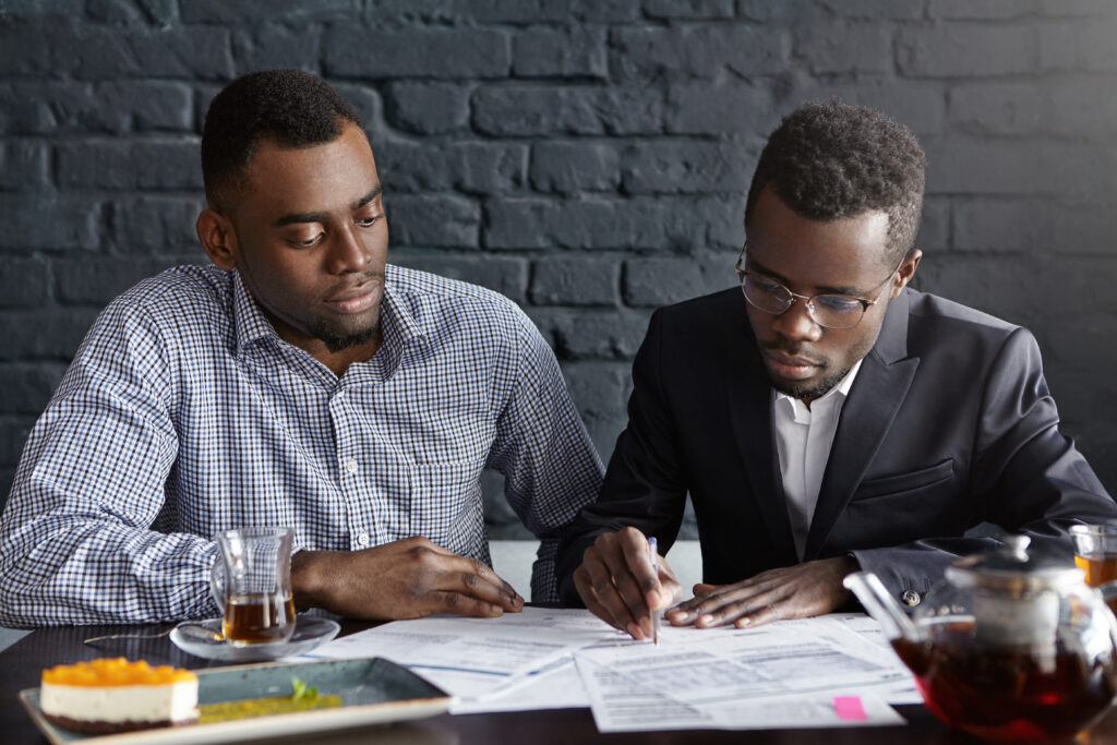 Two confident African-American businessmen dressed formally having serious concentrated looks while signing contract during business meeting at cafe, man with pen showing his partner where to sign